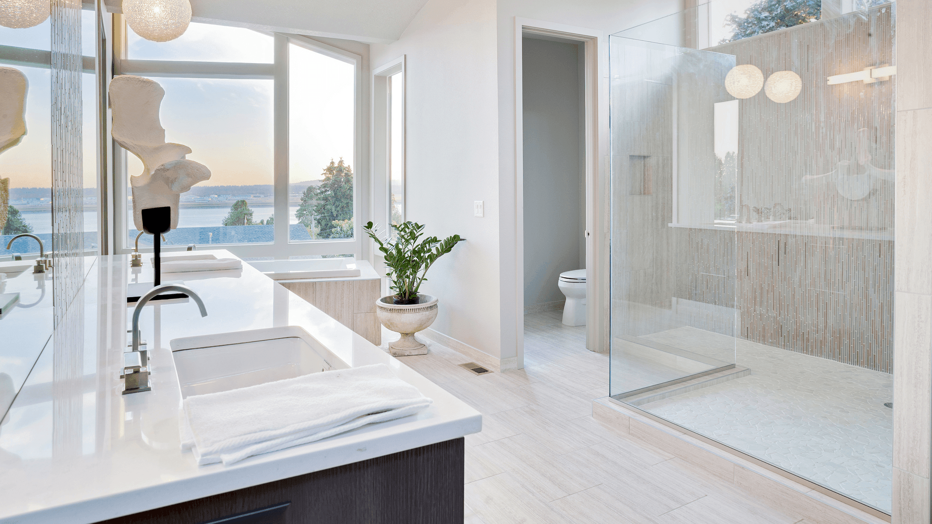 Modern bathroom with a large shower, white countertops, and a plant under a scenic window view.